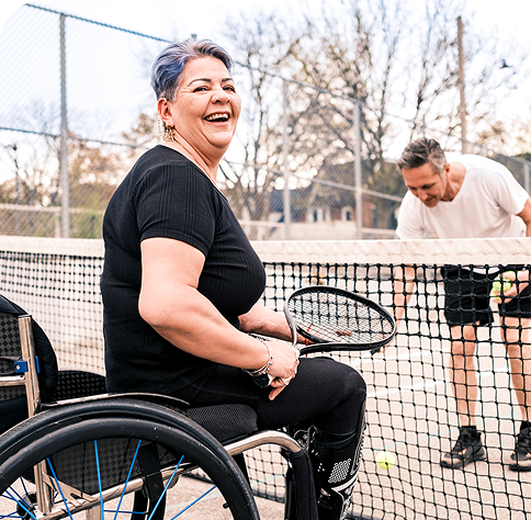 Smiling woman in a wheelchair holding a tennis racket on a tennis court, with a man in the background adjusting the net.